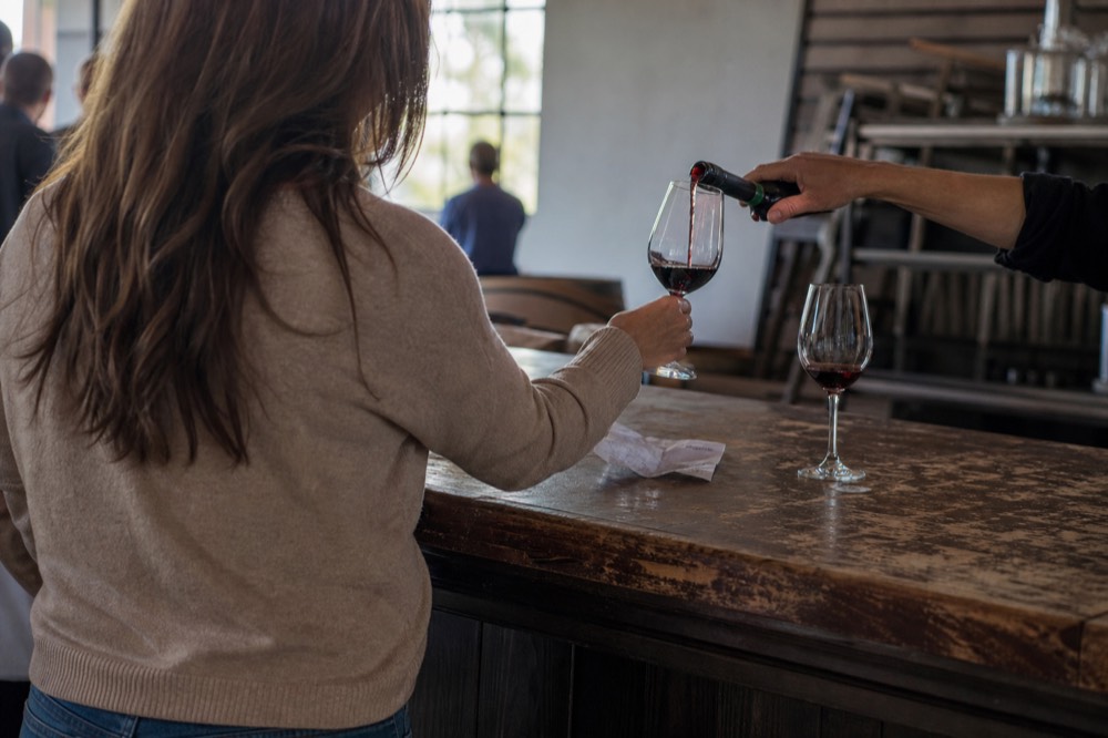 A guest receiving a poured taste of red wine across a tasting room counter